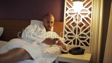一个穿浴袍的男人躺在床上，打电话给酒店的接待处