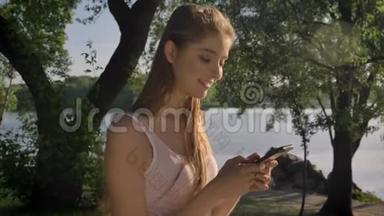 年轻漂亮的女人在她的手机上打字，在<strong>河边</strong>的<strong>公园</strong>里看着镜头，微笑着，美丽的景色