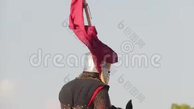在中世纪的锦标赛上，一个身穿盔甲，手里拿着旗子的人骑着一匹马，经过其他穿着历史服装的人