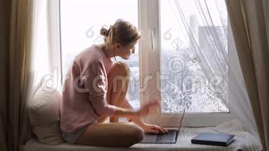 坐在靠近窗户的窗台上的女人正在用她的笔记本电脑工作