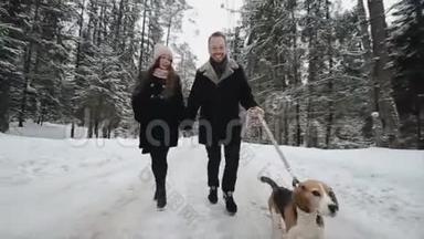 年轻漂亮的家庭在冬天的森林里和一只狗玩。 男人和女人在森林里和比格一起跑步。
