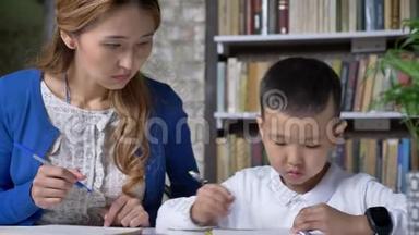 亚洲小男孩和年轻的母亲一起做家庭作业，女人帮助孩子，坐在桌子后面，书架背景