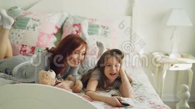 美丽快乐的母亲和小女儿在电视上看卡通电影，在家里躺在床上大笑