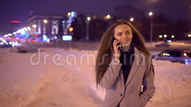 智能手机的女人晚上在街上用智能手机说话。 一位英俊的年轻休闲女士在手机上聊天