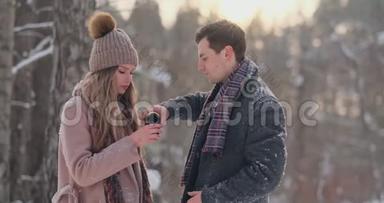情侣在冬天的森林里喝热水瓶里的茶。 冬天公园里穿外套的时髦男女