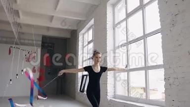漂亮的少女体操运动员在靠近窗户的白色背景的体育体育馆里用彩带跳舞。 吸引力