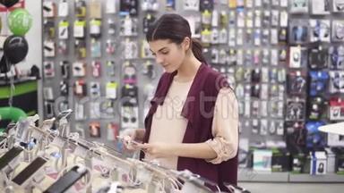年轻美丽的黑发女孩微笑，滚动一个iPhone绑在<strong>展示货架</strong>在电器商店。 选择新的