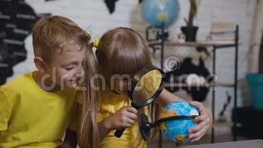 一个漂亮的女孩和一个男孩通过放大镜看着教室里的地球仪。 上地理课