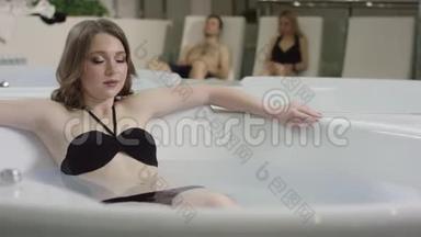 穿着<strong>蓝</strong>色眼睛的黑色泳衣的漂亮女孩，在人们休息的背景下，在按摩浴缸里放松