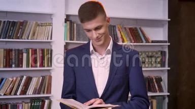 年轻英俊的商人正在看书，看着相机，微笑着，书架在后台