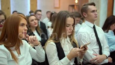 年轻的女学生在观众室内使用智能手机为工作写课文。 从团体后面看