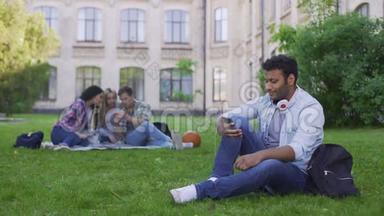 年轻的西班牙裔男学生坐在草地上，在手机上滚动，放松