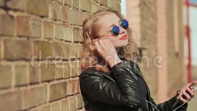 时尚的金发女郎戴着太阳镜和皮夹克，用手机在<strong>蓝牙耳机</strong>上听音乐
