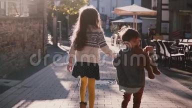 小女孩和男孩牵着手一起走。 慢动作。 后景。 在一个阳光明媚的日子里，两个孩子在老城闲逛。