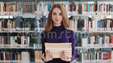 微笑的女学生拿着书站在图书馆的书架前
