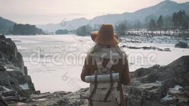 一个年轻的女孩旅行者在智能手机上拍照。 拍摄一条冰冻的冬季河流.. 一顶帽子和一个旅行者`背包