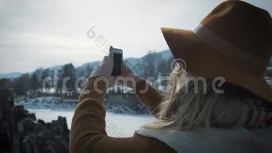 一个年轻的女孩<strong>旅行</strong>者在智能手机上<strong>拍照</strong>。 拍摄一条冰冻的冬季河流.. 一顶帽子和一个<strong>旅行</strong>者`背包