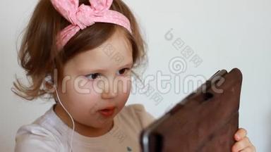 一个戴着耳机的小可爱女孩看着<strong>屏幕</strong>监视器平板电脑。 看电视、录像、<strong>卡通</strong>或玩游戏的滑稽儿童