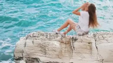 美丽的年轻女孩坐在岩石上，挺直了头发，享受着清爽的微风和美丽的景色。