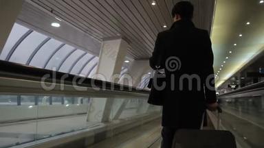 一个穿着黑色外套，带着手提箱的人正在乘坐机场自动扶梯返回。