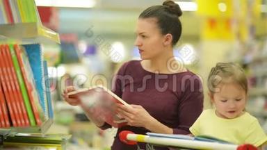 年轻的母亲和她的女儿在超市选书。 美丽的女儿坐在超市的购物车里