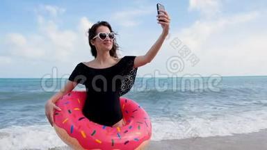 年轻漂亮的女孩穿着泳装微笑，在海滩自拍，海背景。 漂亮的带甜甜圈的时髦女人