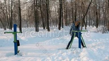 穿着蓝羽绒服带头罩的女人在冬季公园前景的模拟器上做腿部运动