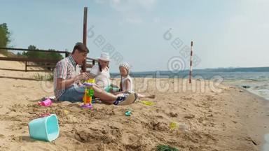 一个穿格子衬衫的成熟男人和两个女孩坐在河边的沙子上。 家庭户外活动