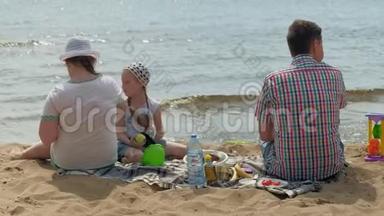 一个穿格子衬衫的成熟男人和两个女孩坐在河边的沙子上。 家庭户外活动