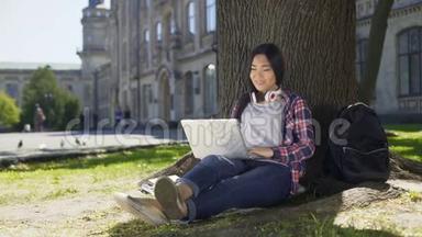 年轻的<strong>大学生</strong>使用笔记本电脑，坐在树下，<strong>微笑</strong>着社交