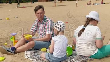 一个穿<strong>格子</strong>衬衫的成熟男人和两个女孩坐在河边的沙子上。 家庭户外<strong>活动</strong>
