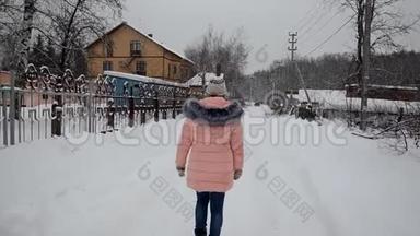 特写黑色靴子在雪地里行走.. 穿着粉红色羽绒服的女孩在白雪覆盖的森林乡间小屋小径上。 冬季