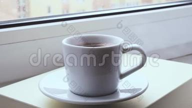 在白雪皑皑的冬日早晨，热气腾腾的咖啡杯站在窗户<strong>上</strong>。 背景下的雪。 女人的手<strong>拿起</strong>一个杯子。