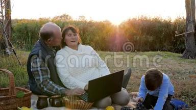 成熟的一对夫妇用笔记本电脑坐在格子花格子上，孙子在日出后野餐