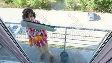 透过窗户观看，微笑的女人戴着耳机，戴着手套，用特殊的拖把清洗窗户，享受她的工作
