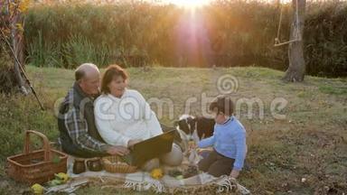 快乐的老人在日出的背景下和孙子、狗坐在格子花格子上，用电脑野餐