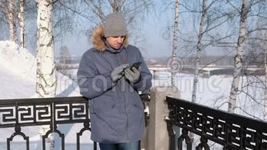 穿着蓝色夹克，带着毛皮罩的人在冬<strong>季</strong>公园里用手戴手套擦拭<strong>手机</strong>屏幕。