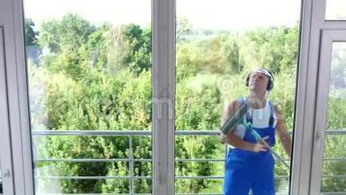 透过窗户观看，英俊的男清洁工人，穿着蓝色工作服，带着耳机，清洁，洗涤
