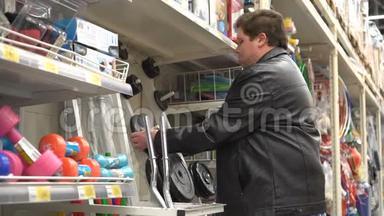 穿黑色皮夹克的胖子检查了超市货架上的杠铃的重量。