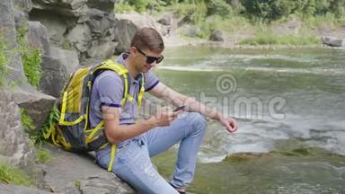 一个带着黄色背包的年轻人坐在一条山河附近。 使用智能<strong>手机</strong>。 旅游及<strong>科技</strong>概念