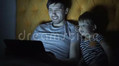 年轻的父亲和他的小儿子晚上躺在家里的床上，用笔记本电脑看卡通电影