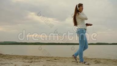 戴着耳机的女孩晚上带着平板电脑在海滩上散步，听音乐。