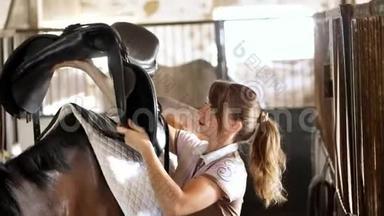 马厩里，一个穿着马服的女骑手在一个棕色的年轻人的背上，为一个马鞍和一个骑马的马鞍做后盾