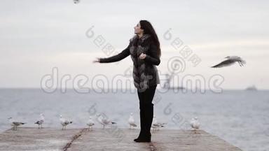 年轻迷人的黑发女人，穿着毛皮<strong>大衣</strong>，在<strong>冬天</strong>在海洋边喂海鸥。 慢镜头