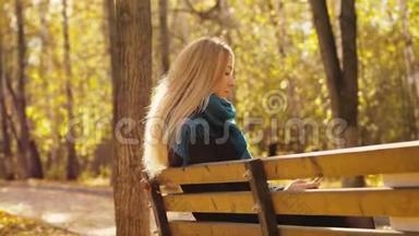 年轻迷人的金发女人坐在长凳上，在秋日公园使用智能手机。 4k，3840x2160
