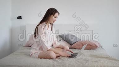 年轻放松的黑发女人白天坐在她家房间的床上，在笔记本电脑上打字