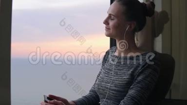 在日落时分，微笑的女孩站在窗边，用手机用耳机听音乐。 好好享受