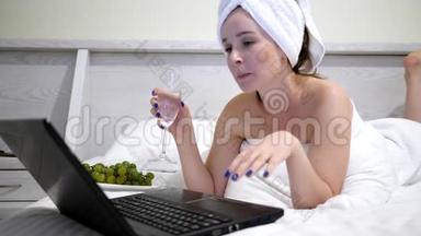 头上戴毛巾的女孩带着香槟在社交网络上交流，在床上用笔记本电脑交流