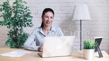 上班时坐在桌旁的一个手提电脑视频通话时，微笑的时髦女孩在说话挥手打招呼