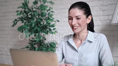 上班时，一个手提电脑坐在桌子旁的视频通话中，微笑的时髦女孩在聊天、挥手和打招呼